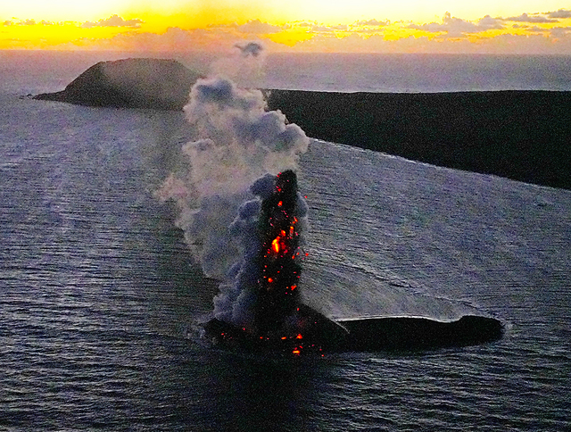 硫黄島の近くで噴火する火山。噴煙に混じって赤く光るマグマを噴き上げていた。奥は摺鉢山＝３日午後５時３２分、東京都小笠原村、本社機から、恵原弘太郎撮影