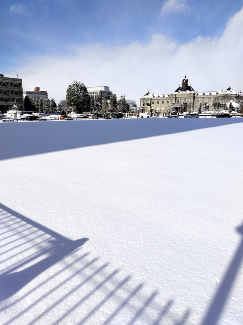 山形市中心部、旧県民会館跡の更地は新雪に覆われ、晴れ間がのぞいた午後には隣のビルなどの影が伸びた=2024年1月16日午後1時26分