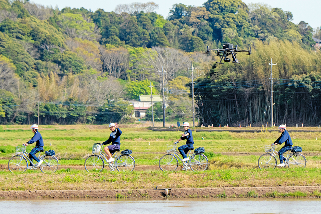 田んぼの中の道を自転車で下校する生徒たち。それをドローンが見守る=2024年4月15日午後2時52分、千葉県東庄町青馬、根岸敦生撮影