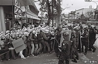 １９７６年１月、沖縄海洋博閉会式に皇太子さまと美智子さまが出席することに反対し、沖縄県本部町の商店の前でデモ行進する新左翼の若者たちと、歩道から車道に進出させまいと盾で規制する機動隊員たち。右端は機動