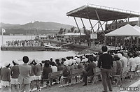 水上のステージに参加各国の国旗を持った警備員が並ぶなか行われた海洋博の閉会式と、式典を見つめるコンパニオンや関係者たち