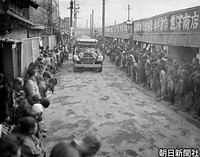 空襲で焼け再建された神奈川区大口町商店街を、多くの住民が頭を下げて迎えるなか、徐行で進む昭和天皇の車列。道路に舗装はなく排水用の溝がある