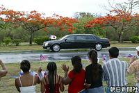 天皇、皇后両陛下がサイパン島の空港から宿舎に向かう車列の沿道に集まり、日の丸、星条旗、北マリアナ自治政府旗を振る住民。真ん中の車窓から皇后さまが手を振られている。後方でオレンジ色の花を咲かせているのは